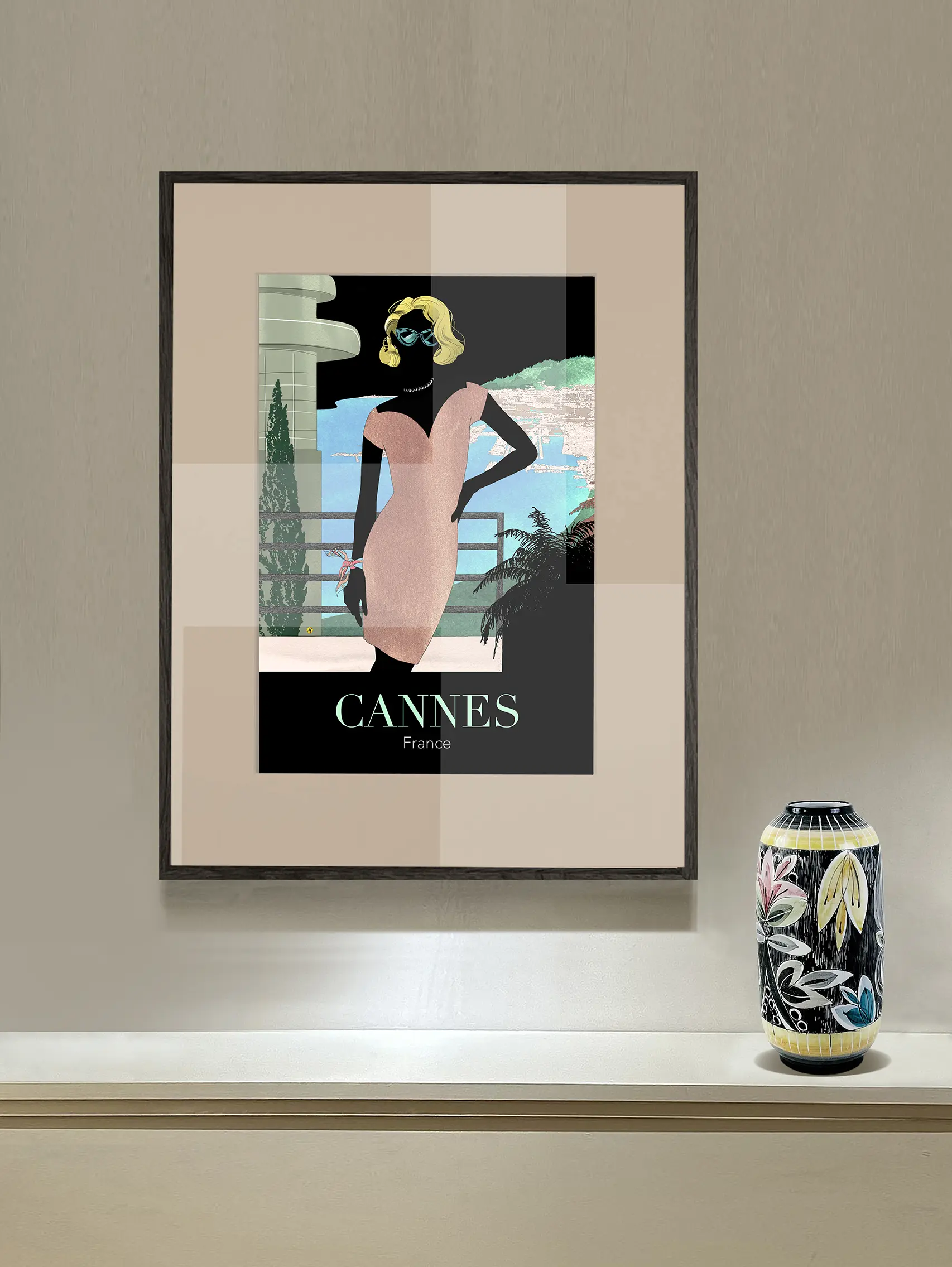 Black frame with a poster showing a black silhouetted woman with yellow wavy hair and green sunglasses. She is wearing a tight light pink dress and a scarf is tied around her wrist. Behind her is a part of an art deco house with a railing and further back is a view in light blue, beige and green of the French riviera city of Cannes. The background is in black. At the bottom you can read the text "Cannes France". Underneath the poster is a shelf with a flowered printed vase in black, yellow, pink and blue. The wall is colored in beige.